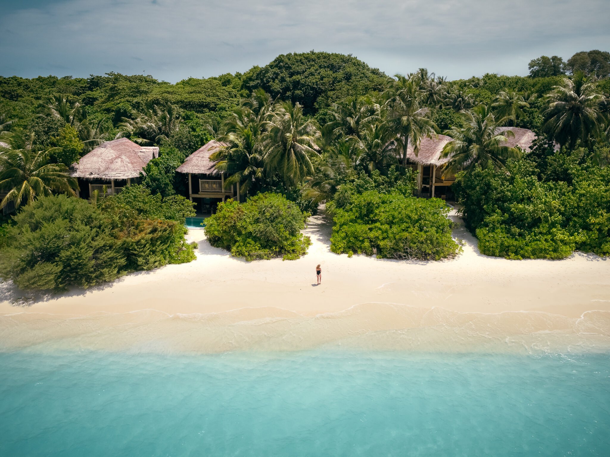 A serene tropical beach features turquoise water, pristine white sand, and lush greenery. Several thatched-roof huts nestle among the palm trees in the background. A single person stands on the beach facing the ocean, enhancing the feeling of tranquility reminiscent of a Maldives luxury resort like Soneva Fushi Villas.